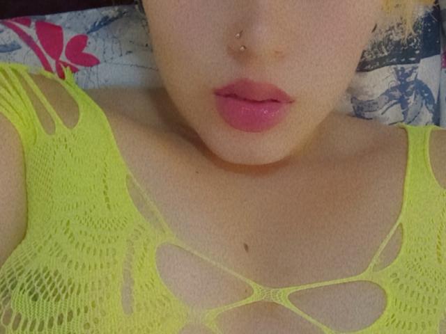 Keidy69's face, with pink lips and a nose piercing, wearing a neon yellow mesh top showing her small tits and a few moles on her chest.