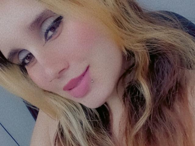 Keidy69 has blonde and brown ombre hair, a lip piercing, pink lipstick, and is smiling at the camera with dark eyeliner. Her tits are not visible.