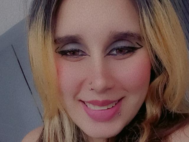 Keidy69 has blonde and dark hair, a nose piercing, a lower lip piercing, pink lipstick, and dramatic eyeliner. She's smiling with pink cheeks.