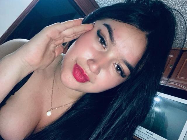 AliceMillerlu with dark hair, red lipstick, and a gold necklace, touching her cheek, showing her tits, and a slight smile in a selfie. Her eyes are half-open.