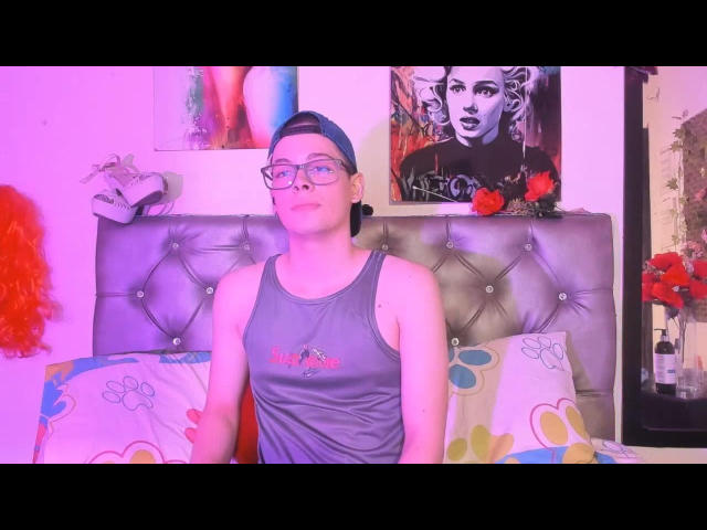 LukaSmith, mit einer blauen Mütze und Brille, sitzt auf einem Bett in einem grauen Tanktop und zeigt eine schlanke Figur mit kleinen Brüsten. Schlafzimmer mit bunten Kissen und Kunstwerken..