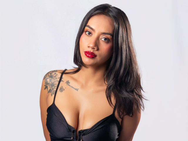 LunaGomezz mit schwarzen Haaren, rotem Lippenstift und einem schwarzen BH, die ihre Brüste zeigt und ein Blumen-Tattoo an ihrer rechten Schulter hat. Sie schaut in die Kamera..