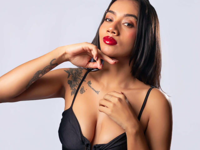 LunaGomezz with red lipstick, black dress, and tattoos on her right shoulder, touching her chin with her left hand, showing her tits and a small tattoo above her chest.