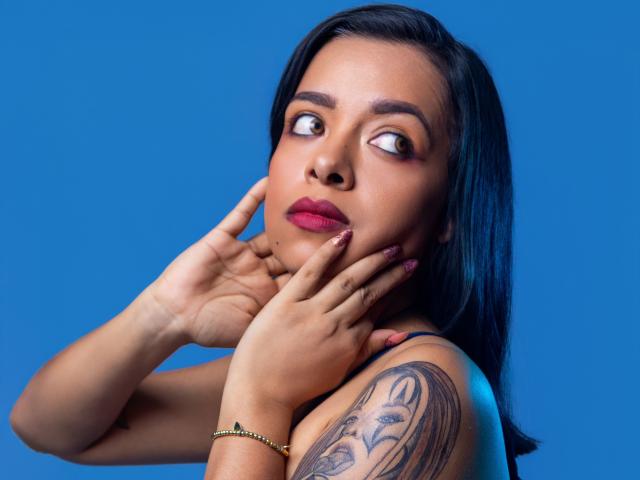 ArielHot69 has dark hair, brown eyes, and red lipstick, with hands near her face, showing a tattoo on her left shoulder, wearing a gold bracelet, against a blue background.
