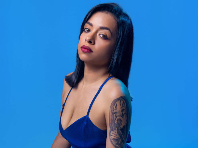 ArielHot69 with black hair, blue dress, red lipstick, and a tattoo of a woman's face on her arm, looking slightly to the side against a bright blue background.