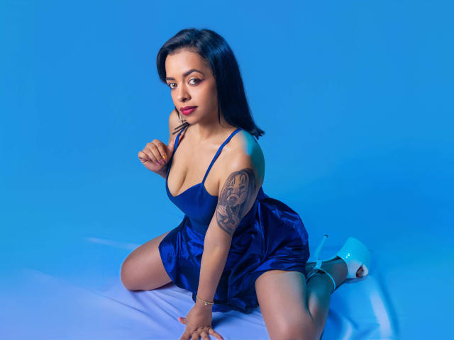 ArielHot69 with dark hair, blue dress, and tattoo on right arm, kneeling, showing tits, in high heels, on blue background. Her hand touches her chin.
