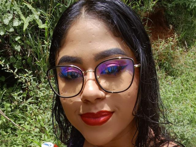 Een vrouw met donker, nat haar, rode lipstick en glanzende paars-blauwe oogschaduw, die een bril draagt, in een groene, buitenomgeving. Haar neus is licht zichtbaar tussen haar oogschaduw en bril..