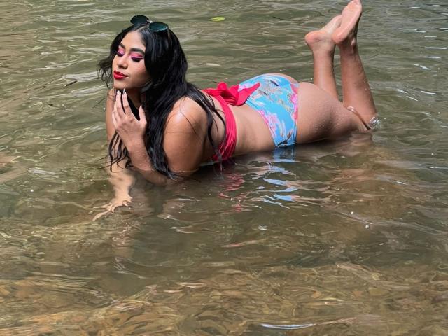 KatikaRouse lying in shallow water, wearing a pink bikini top and blue patterned bottoms, her ass up, black hair, red lipstick, sunglasses on head, hands near face, legs up.