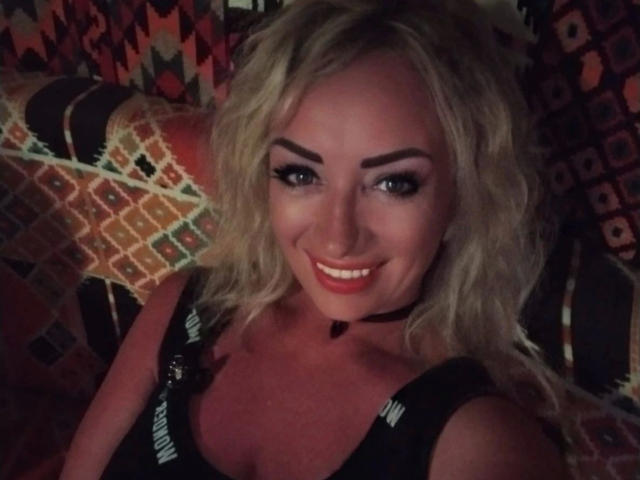 MilfPamela: bellissima, con capelli biondi ondulati, sopracciglia folte, rossetto rosso, collana nera, sorride alla telecamera, indossa una maglietta nera con spalline, mostrando il seno..