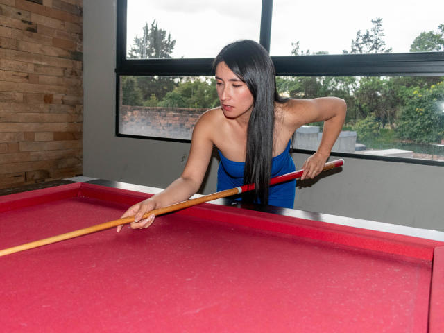 JainaRogers, with long black hair, blue dress, and tan skin, bends over a red pool table, aiming a wooden cue stick at a ball.