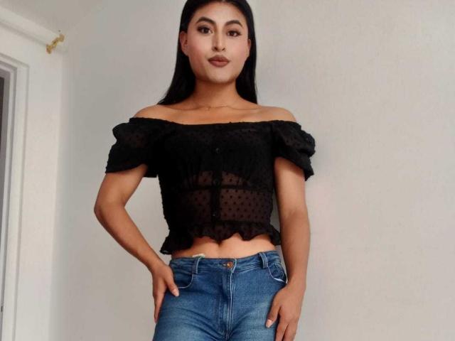 MichelleDenise has tan skin, black hair, and is wearing a sheer black off-shoulder top with polka dots and blue jeans, standing with hands on her hips.