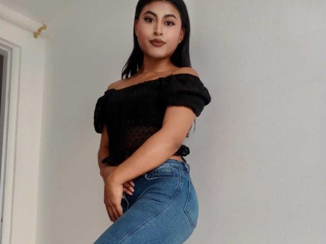 MichelleDenise with black off-shoulder top, small tits, medium ass, wearing blue jeans, standing, looking at camera, hands on hip, light skin, long black hair, smiling.