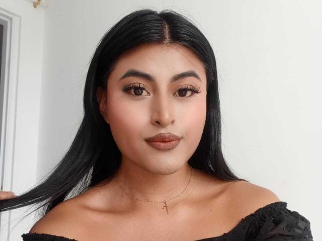 MichelleDenise has long black hair, brown eyes, and wears a black off-shoulder top with a cross necklace. Her makeup includes dark lipstick and subtle eyeliner.