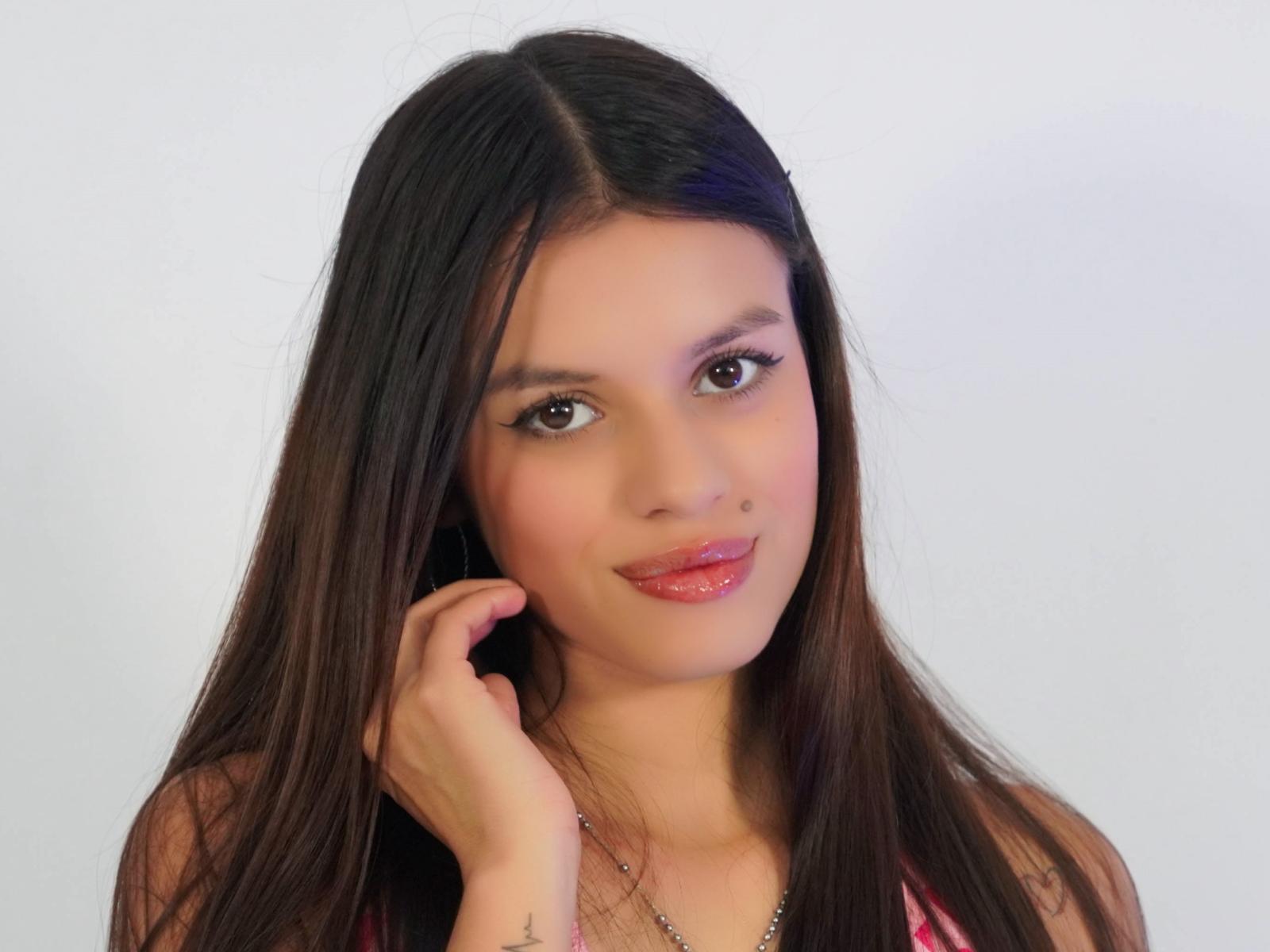 LunaVera has long brown hair, brown eyes, glossy pink lips, and a cute smile, with her hand touching her cheek, wearing a pink top, looking sexy.