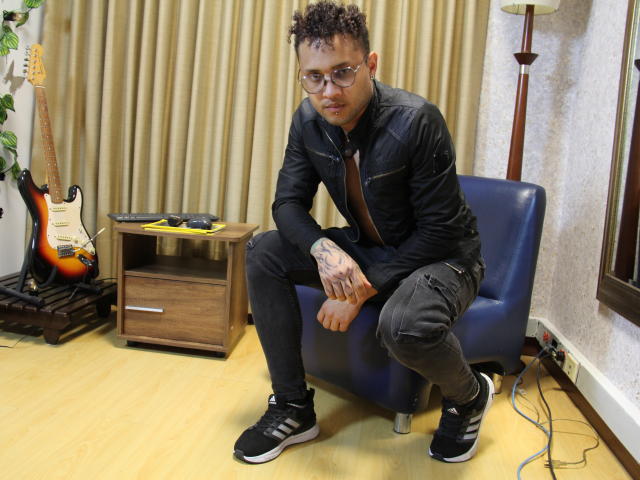 OrfeoJames, short curly brown hair, glasses, black jacket, gray pants, black sneakers, sitting on blue chair, tattooed hand, guitar and wooden table in background.