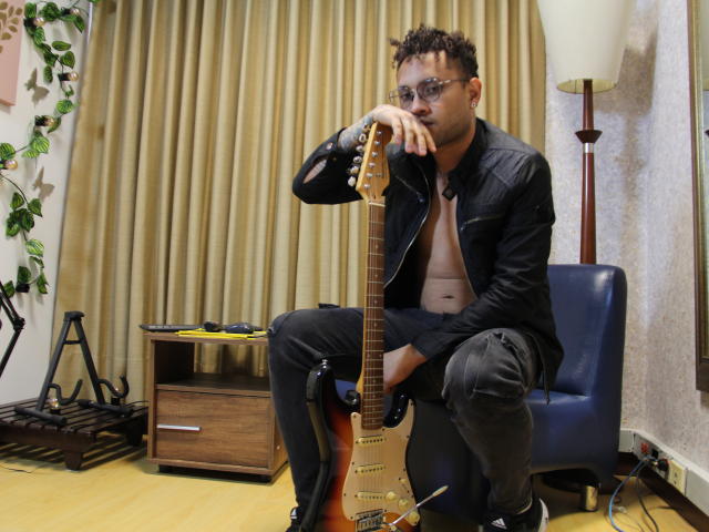 OrfeoJames, with curly hair and glasses, sits shirtless, holding a sunburst electric guitar, wearing black pants and a leather jacket, in a room with a blue chair.