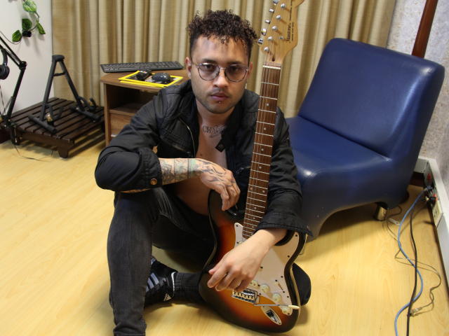 OrfeoJames with short curly hair, glasses, black jacket, tattoos, sitting on floor holding sunburst electric guitar, blue chair behind, wooden floor, and music equipment in background.