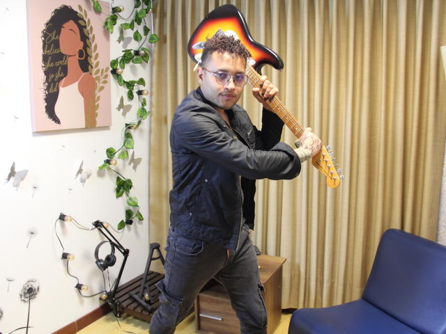 OrfeoJames, wearing glasses and a black jacket, holding a sunburst electric guitar, standing with a rock pose, curly hair, and a slightly messy mustache, in a room.