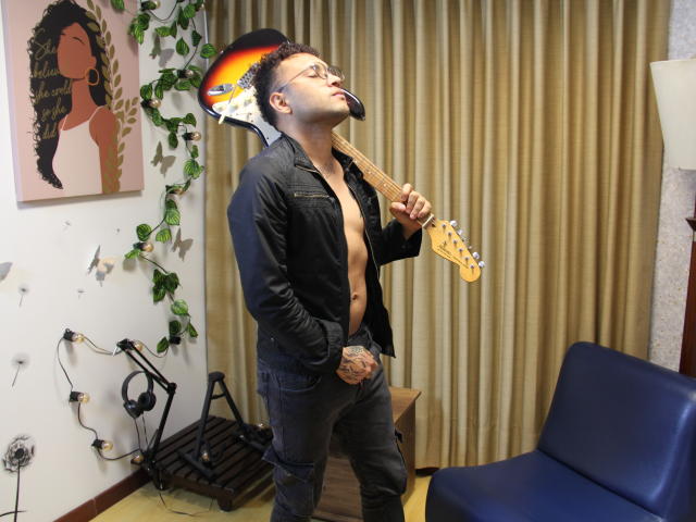 OrfeoJames, shirtless with black pants, holding a sunburst electric guitar, tongue out, leaning back, curly black hair, glasses, tattoos on hands, standing in a room with a blue chair.