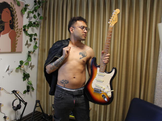 OrfeoJames with short curly hair, glasses, tattoos, and a leather jacket, holding a sunburst electric guitar, wearing black pants with a visible "Pussy" tattoo on his lower belly.