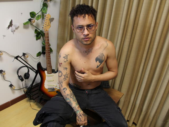 Shirtless OrfeoJames with tattoos, glasses, and curly hair, sitting on a bench, hand on chest, wearing black pants, guitar and headphones in background, beige curtains behind.