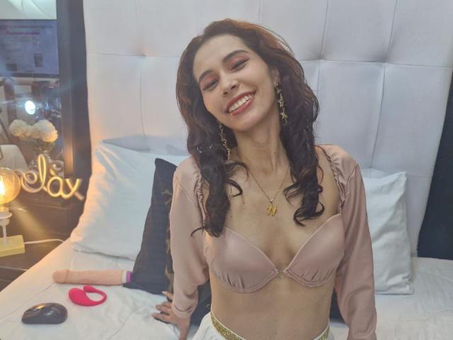 KatheKiss with wavy brown hair, smiling, wearing a pink bra and long-sleeve top, gold necklace, and earrings, sitting on a bed with a dildo and vibrator.
