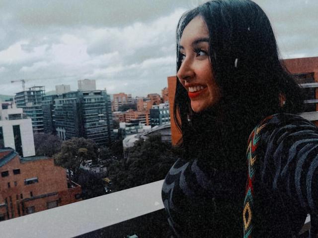 SusanMonroy with medium tits, smiling, black hair, wearing a dark sweater with colorful patterns, standing on a balcony, looking to the side, city skyline in the background.