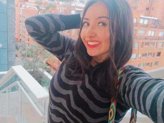 SusanMonroy with wavy black hair, smiling, wearing a black striped sweater, nose ring, and colorful shoulder bag, standing on a balcony, hand in hair.