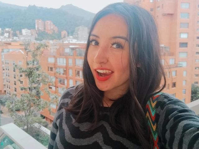 SusanMonroy with long dark brown hair, wearing a black zigzag sweater, smiling with red lipstick, showing her teeth, standing on a balcony with a city and hills background.