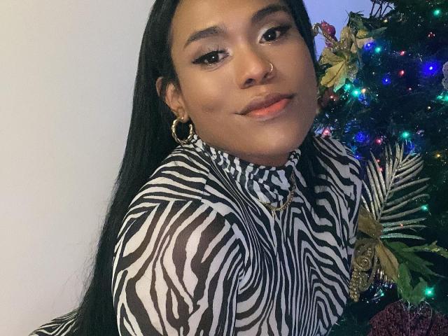 AshleyRob with long black hair, zebra-pattern dress, gold hoop earrings, nose ring, and subtle makeup, standing next to a decorated Christmas tree with colorful lights.