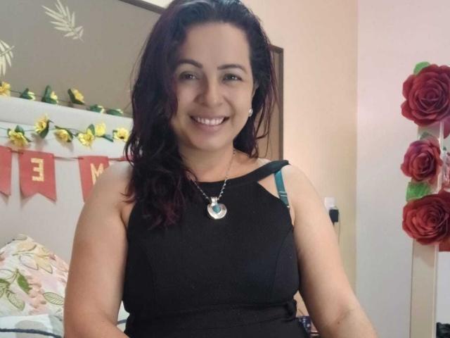 PamelaSmiths tiene el cabello castaño oscuro, está sonriendo y lleva una camiseta negra sin mangas con un collar de plata, mostrando sus senos y una banda negra. Está de pie en un dormitorio..