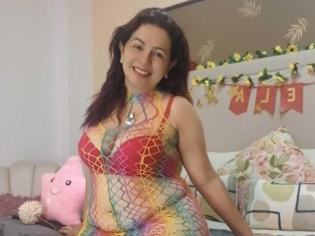 Pamela Smiths trägt ein farbenfrohes Netzhemdkleid, einen roten BH und ist oben ohne, lächelt, hat dunkles Haar, ein pinkes Kissen, Blumenkissen, gelbe Blumen und im Hintergrund ein "Happy"-Banner..