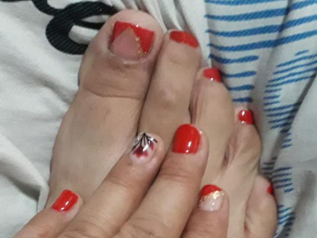 Les orteils de PamelaSmith sont vernis d'un rouge brillant, dont l'un présente un motif blanc et argenté, et sa main les tient doucement ensemble. Sa peau est claire et ses ongles sont brillants..