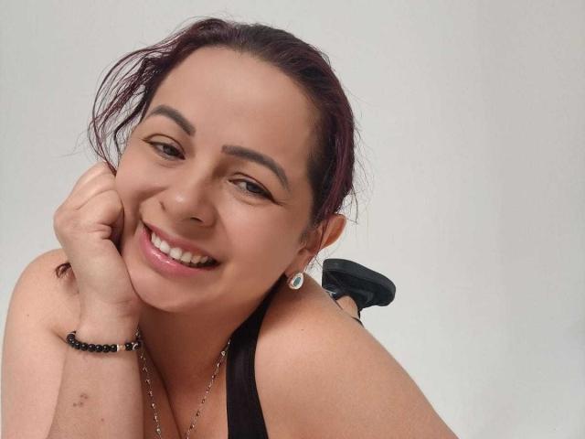PamelaSmiths heeft donkerrood haar, glimlacht, toont haar tanden, draagt een zwarte tanktop en heeft een zwarte armband om haar rechterpols, met een witte achtergrond..