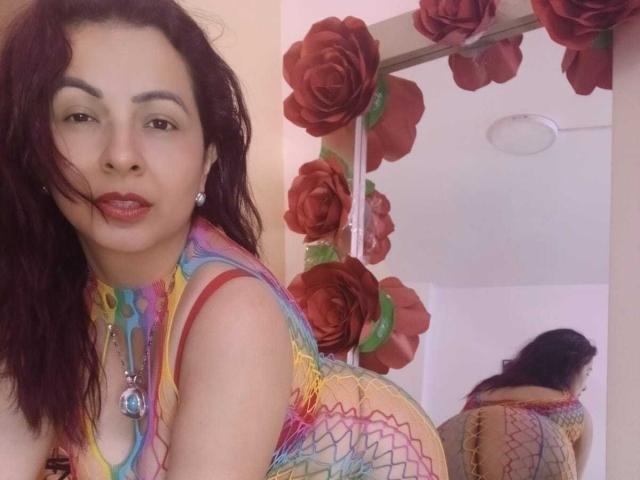 Pamela Smiths, met lang donker haar, draagt een kleurrijke, netachtige top, rode lippenstift en een blauwe ketting, en staat voor een spiegel met rode rozen decoraties..