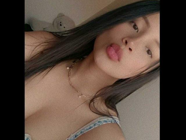 TinyLaurent a des lèvres roses et fines, de longs cheveux noirs, et porte un collier fin avec de petites perles. Elle porte un débardeur bleu, qui révèle ses seins..