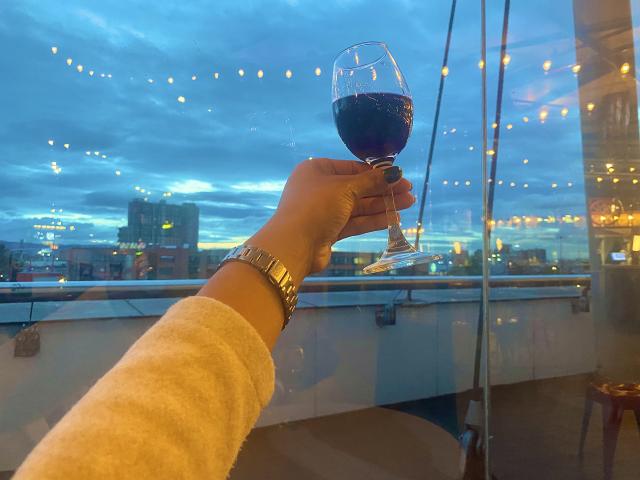 SophieSonyas' hand with a gold watch, holding a glass of red wine, her arm in a beige sweater, against a city skyline at dusk, with string lights.