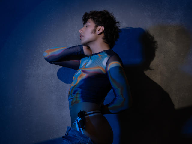 TonyDan, che indossa una camicia a righe colorata a maniche lunghe, ha una mano dietro la testa, in piedi con i glutei leggermente scoperti, i jeans blu parzialmente abbassati, illuminato da una luce blu..