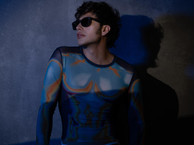 TonyDan mit lockigem, dunklem Haar, der schwarze Sonnenbrillen trägt und ein blaues, orangefarbenes und schwarzes abstraktes Langarmshirt, steht vor einer strukturierten Wand und wirft einen Schatten..