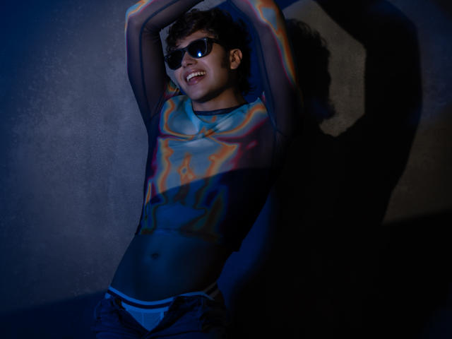 TonyDan con vernice colorata sul petto, braccia alzate, che indossa occhiali da sole neri, sorridendo, biancheria intima blu, jeans scuri e un torso dipinto, in piedi contro un muro in ombra..