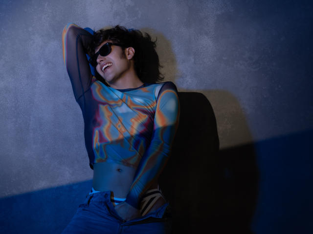 TonyDan, con i capelli neri ricci, indossa occhiali da sole, una maglia a maniche lunghe colorata e jeans blu, è appoggiato a un muro, sorridendo, con una mano dietro la testa, mostrando il sedere..