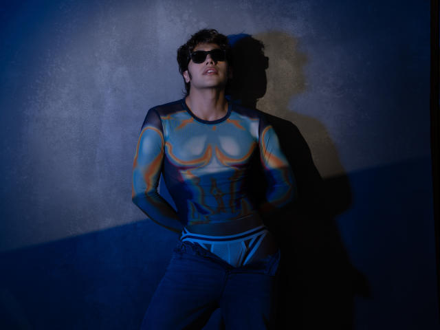 TonyDan mit lockigem, braunem Haar, der schwarze Sonnenbrillen, ein farbenfrohes, halbtransparentes Langarmshirt und blaue Jeans trägt, steht vor einer dunklen Wand und wirft einen Schatten..