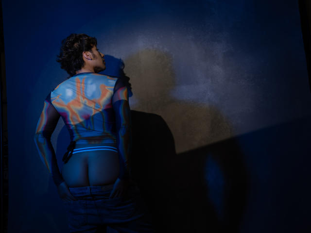TonyDan, le dos peint, les fesses exposées, portant un jean déboutonné, debout contre un mur sombre, une ombre sur le mur, des motifs géométriques bleus et orange sur le dos, cheveux bouclés, regardant vers le haut..