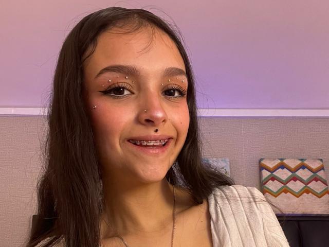 MarietteJones has a cute smile with small nose piercings, dark eyeliner, and a lip piercing. She's wearing a white top with long brown hair. Her ass is not visible.