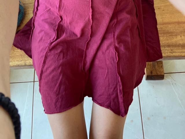 Tapira-hot's pink skirt, showing her ass, with visible creases, standing on white tiles, a wooden table in the background, and a black bracelet on her left wrist.
