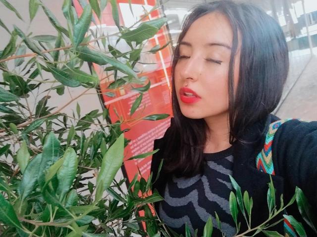 SusanMonroy with her eyes closed, red lipstick, and black zigzag top, standing next to green bamboo leaves, wearing a colorful shoulder bag, has medium-length dark hair.