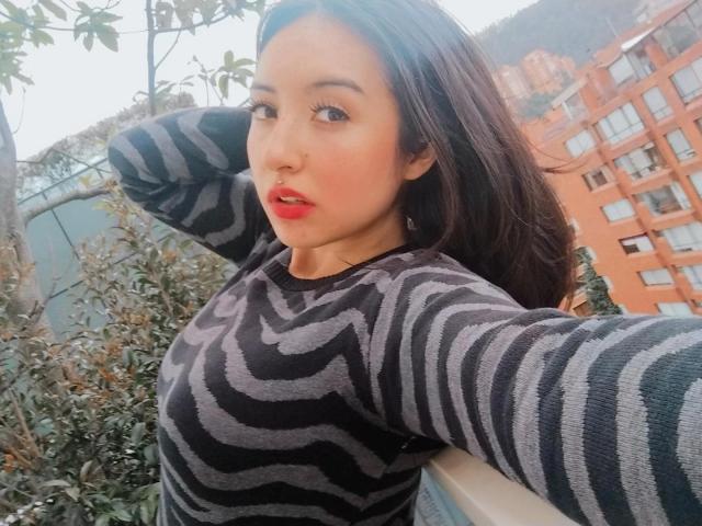 SusanMonroy with red lips, black and gray striped sweater, and medium tits, posing with arms behind head, dark hair, and slight blush, on a rooftop with leafy plants.