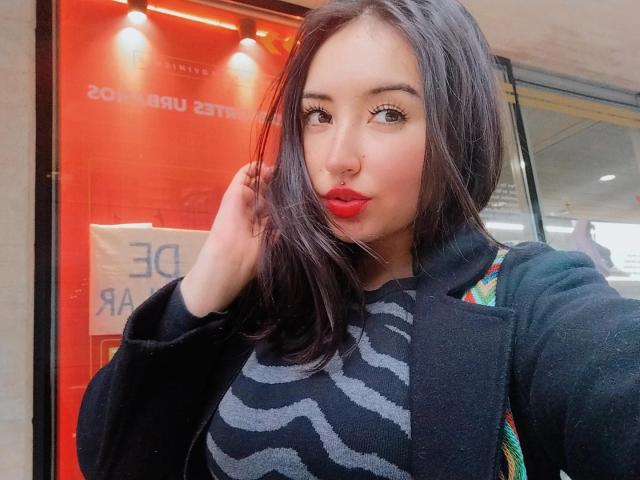 SusanMonroy has medium tits, red lipstick, and black wavy hair. She's wearing a black zigzag pattern top and a black jacket, with a colorful bag strap.