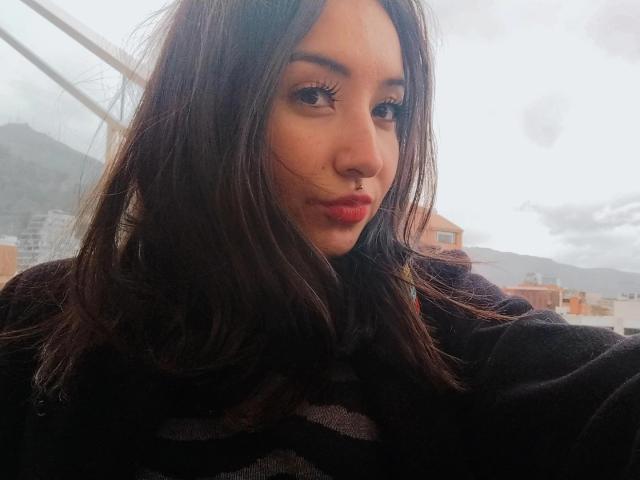 SusanMonroy with long brown hair, red lipstick, black sweater, nose ring, and dark eyelashes, looking at camera, cloudy sky, and mountain in background.