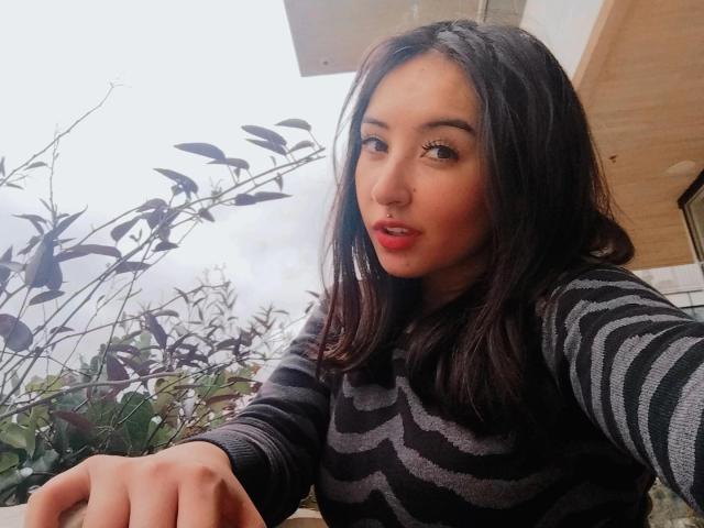 SusanMonroy with dark brown hair, wearing a black and gray striped sweater, has red lipstick, pouty lips, and a cute nose, looking at the camera with a slightly open mouth.